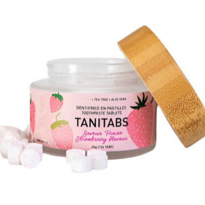 Glass jar of TANITABS Strawberry Toothpaste Tablets with bamboo lid and scattered pink chewable tablets on a white background