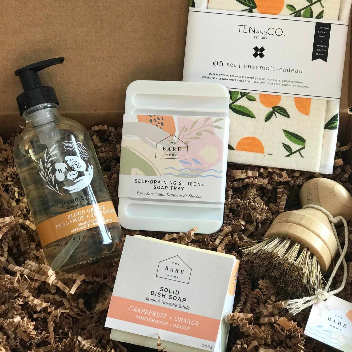 This made in Canada The Bare Home zero waste gift set features a TEN and CO. sponge cloth and tea towel set in a citrus pattern, a refillable 236 ml blood orange, bergamot and sandalwood hand soap in glass bottle, a white silicone soap dish, a grapefruit and orange dish soap bar and a pot scrubber brush
