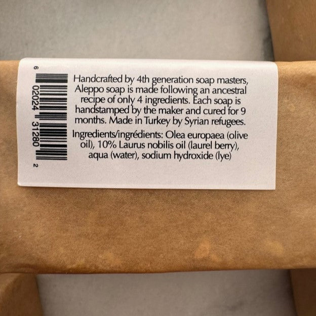 Close-up of the Aleppo Soap ingredients label, detailing its traditional four-ingredient formula of olive oil, 10% laurel berry oil, water, and lye. Handcrafted by 4th-generation soap makers and cured for 9 months in Turkey by Syrian refugees.