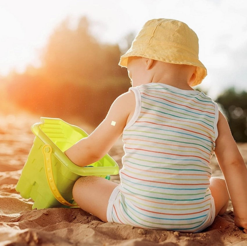 Baby in a striped outfit and yellow hat playing in the sand with a green bucket.