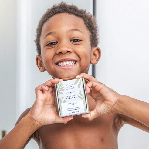 A smiling young boy holds a box containing the bergamot lime hand and body bar soap made in Canada by The Bare Home.