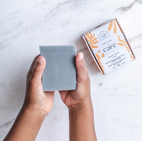 A child's hand holds a bar of 100 g blood orange, bergamot and sandalwood hand and body soap beside the decorative box.