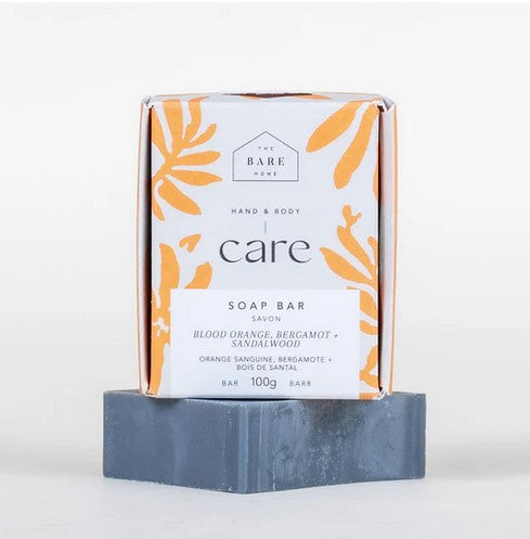 The 100 g charcoal coloured blood orange bergamot and sandalwood hand and body bar soap is pictured on a white background with the soap bar box on top of it in white and orange.