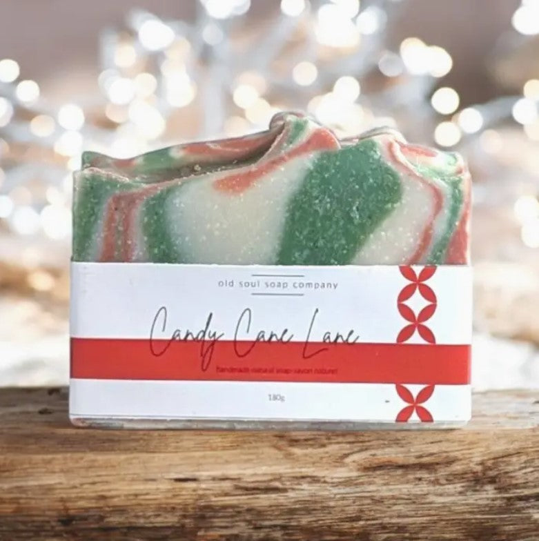 Red and white labelled Candy Cane Lane Canadian made Old Soul Soap Company bar soap with red and green swirls with a candy cane peppermint scent on a wood table with white lights in the background.