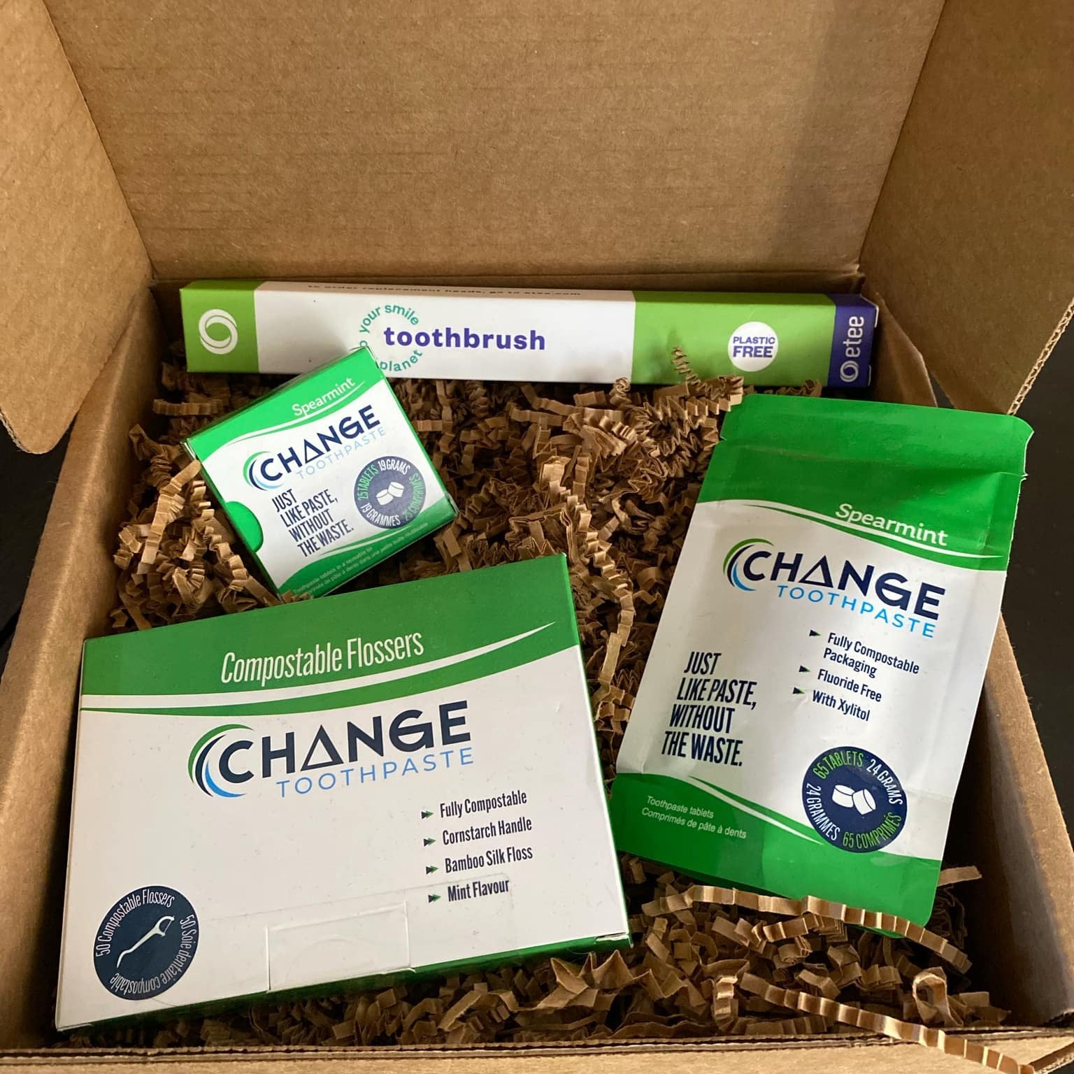 This dental care starter kit features three products from Change toothpaste. A package of 50 compostable flossers, a 65 tablet pouch of spearmint toothpaste tablets, a tin of 25 spearmint toothpaste tablets and a bamboo toothbrush with a replaceable head free etee. These items are shown in an open gift box on top of brown crinkle paper packaging.