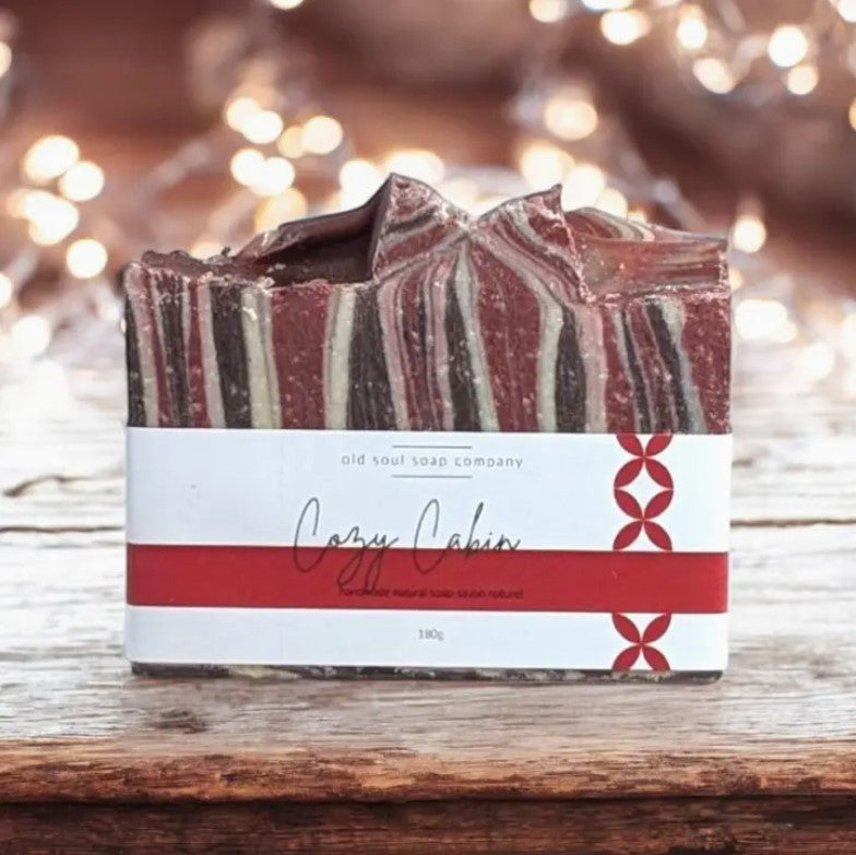 Red and white labelled Cozy Cabin Canadian made Old Soul Soap Company bar soap with red, white and dark brown swirls with an invigorating cedarwood spearmint scent sits on a wood table with white lights in the background.