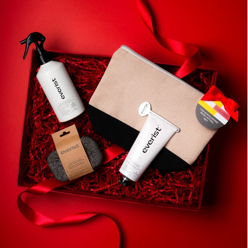 For holiday display purposes a full-size Exfoliating Body Wash Concentrate, Body Oil Spritz, Exfoliating Konjac Body Sponge and a Black Travel Bag are displayed in a red box with red crinkle paper and red ribbon on a red background.
