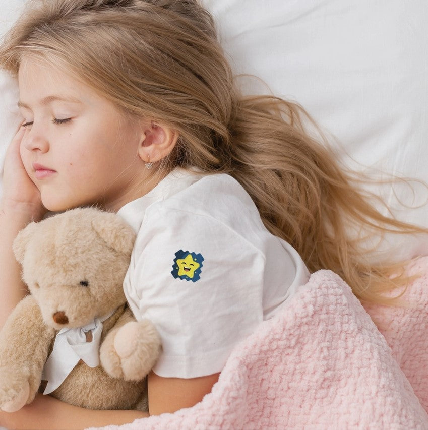 Child sleeping with a teddy bear and pink blanket, wearing a white shirt with a bedtime sticker on the sleeve