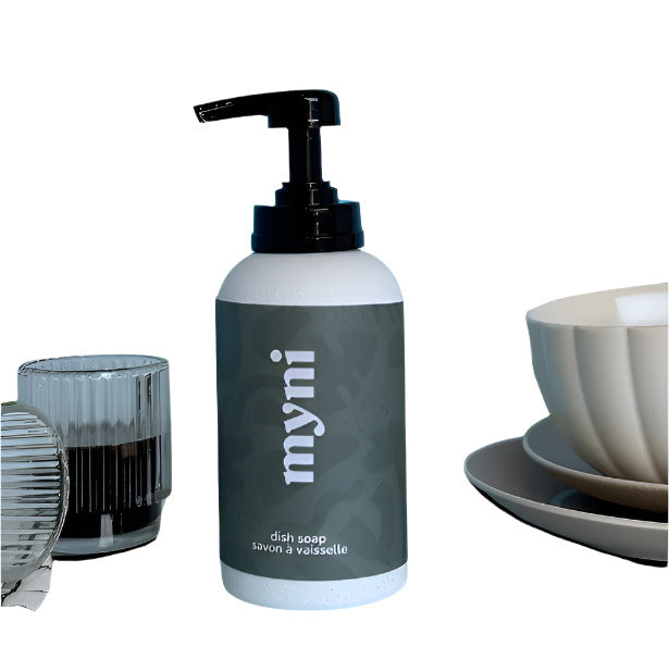 Myni's non-toxic dish soap in a special edition compostable wheat straw bottle with black wrap and white lettering on an otherwise white bottle. The black pump top bottle is pictured beside some dishes against a white backdrop.