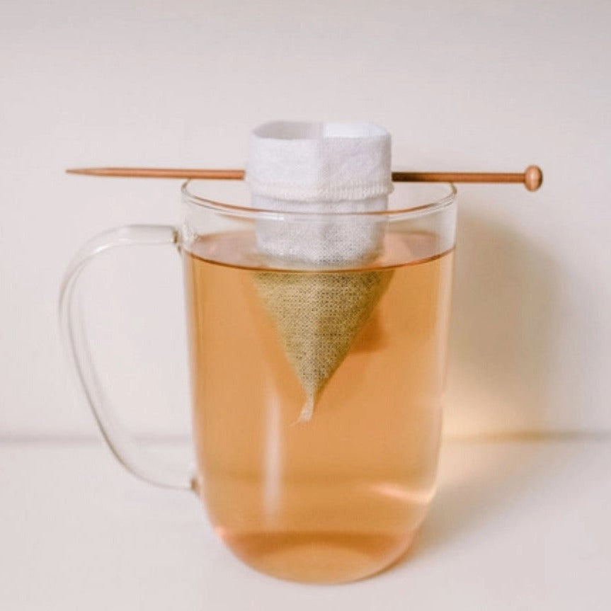 Organic cotton reusable tea bag steeping loose leaf tea in a clear glass mug with wooden balancing stick – plastic-free sustainable tea accessory made in Canada.