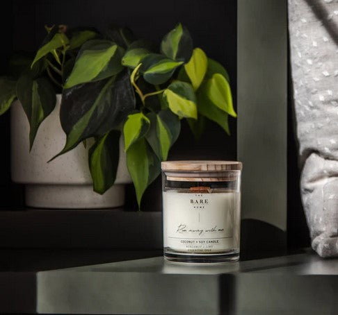 A lit 'Run away with me' candle in a jar sits on a table with a pillow on one side and a plant in the background.
