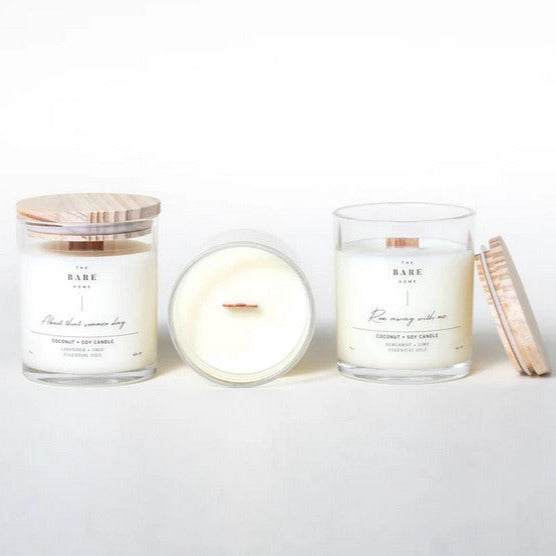 Three candles by The Bare Home in clear glass jars sit in a row with wood tops on a white background. The first one has the lid on, the middle one in laying on its side to show the wooden wick and the last one has the lid off with the lid beside it.