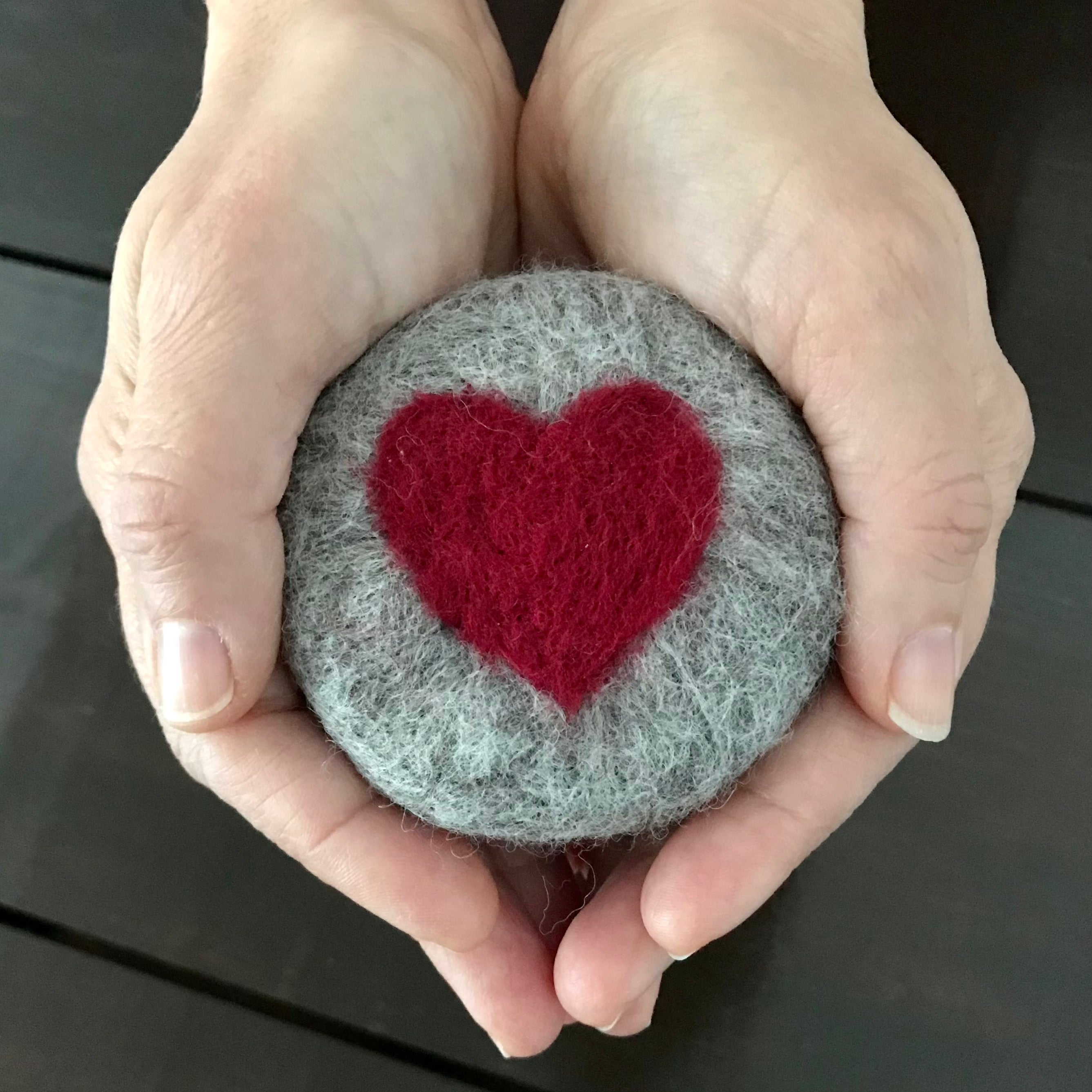 A round hand felted rosemary mint essential oil soap made in Canada in 100% charcoal grey wool with a red heart on the top sits in a woman's palm