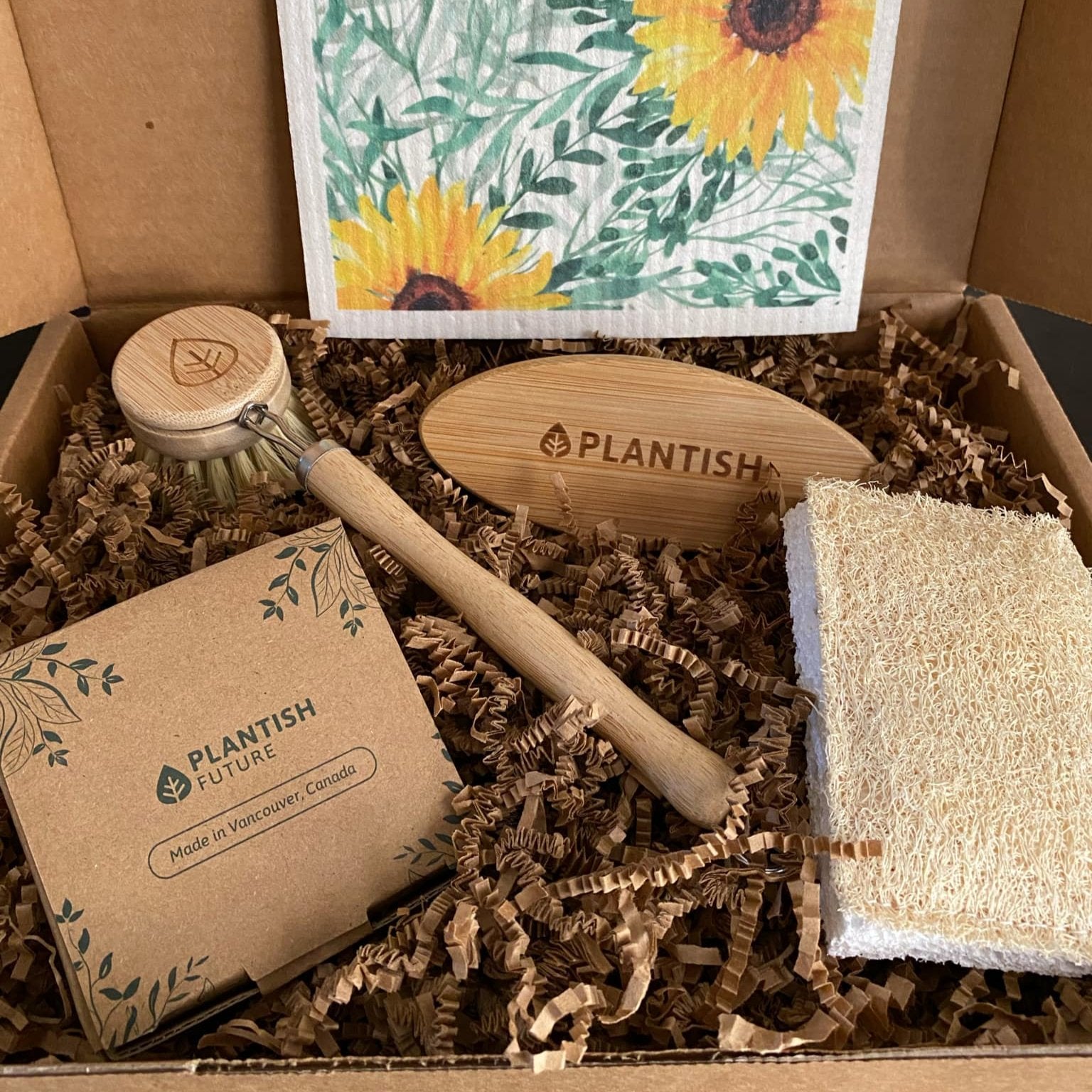 This Sunflower Kitchen Gift Box featuring a Swedish sponge cloth with a sunflower motif, dish soap cake in an aluminum tin, replaceable head dish brush, vegetable brush, and eco scrubber sponge is shown in an open gift box.