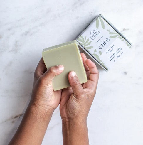A child's hand holds a bar of 100 g bergamot lime hand and body soap beside the decorative box.