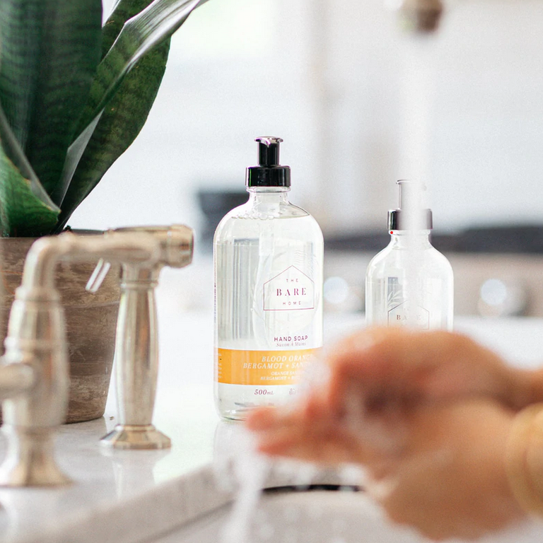 Canadian made The Bare Home Blood Orange, Bergamot Lime and Sandalwood essential oil scented natural hand soap in a 476 ml and 236 ml refillable glass pump bottles sit beside a sink where someone is washing their hands