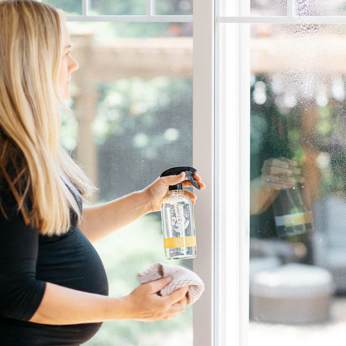 Canadian made lemon tea tree essential oil scented natural all purpose cleaner in a 476 ml refillable glass spray bottle made by The Bare Home company is being used by a pregnant woman on a window