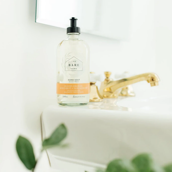 Canadian made The Bare Home Blood Orange, Bergamot Lime and Sandalwood essential oil scented natural hand soap in a 476 ml refillable glass pump bottle sits beside a polished bathroom faucet with plants in the foreground