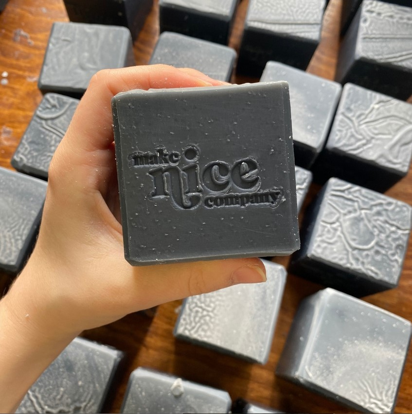 Square solid dish soap cube made with the Make Nice Company logo imprinted on the top is held in one hand with a collection of other dish soap bars in the background