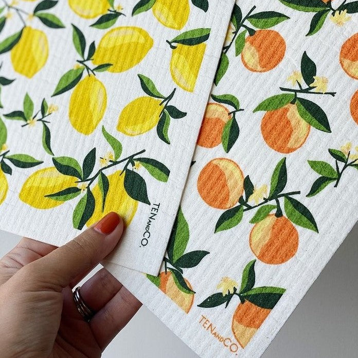 A hand holding pair of Ten and Co. Swedish dishcloths one with a lemon pattern and one in an orange pattern both with leaves and on white backgrounds