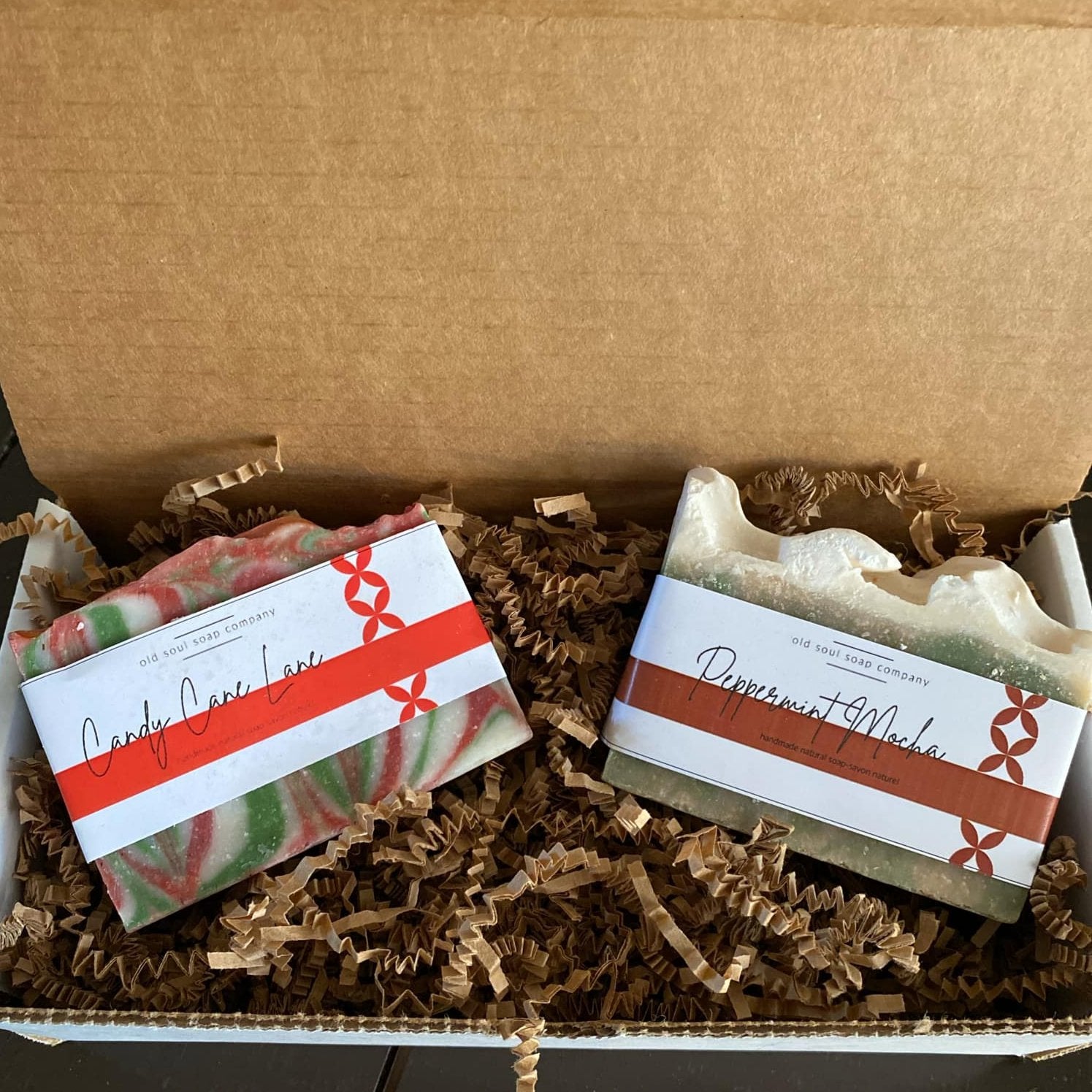 Two labelled handcrafted Old Soul Soap Company 'Candy Cane Lane' and 'Peppermint Mocha' bar soaps in an open white gift box sitting on brown crinkle paper packing. 