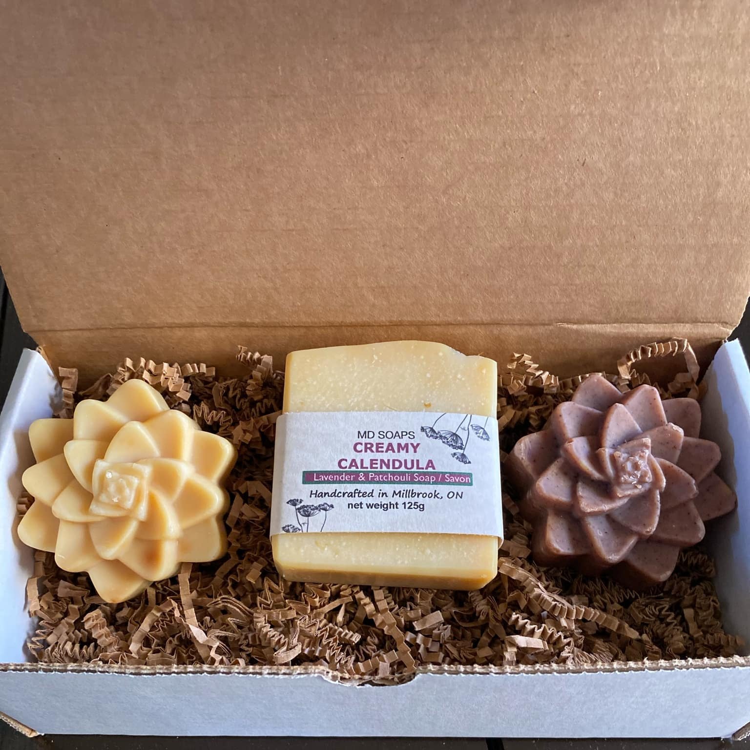 This MD Soaps Gift Set features three lightly fragrant handcrafted soaps including two succulent shaped and on either side of a bar of calendula soap and is pictured on brown crinkle paper in an open gift box.