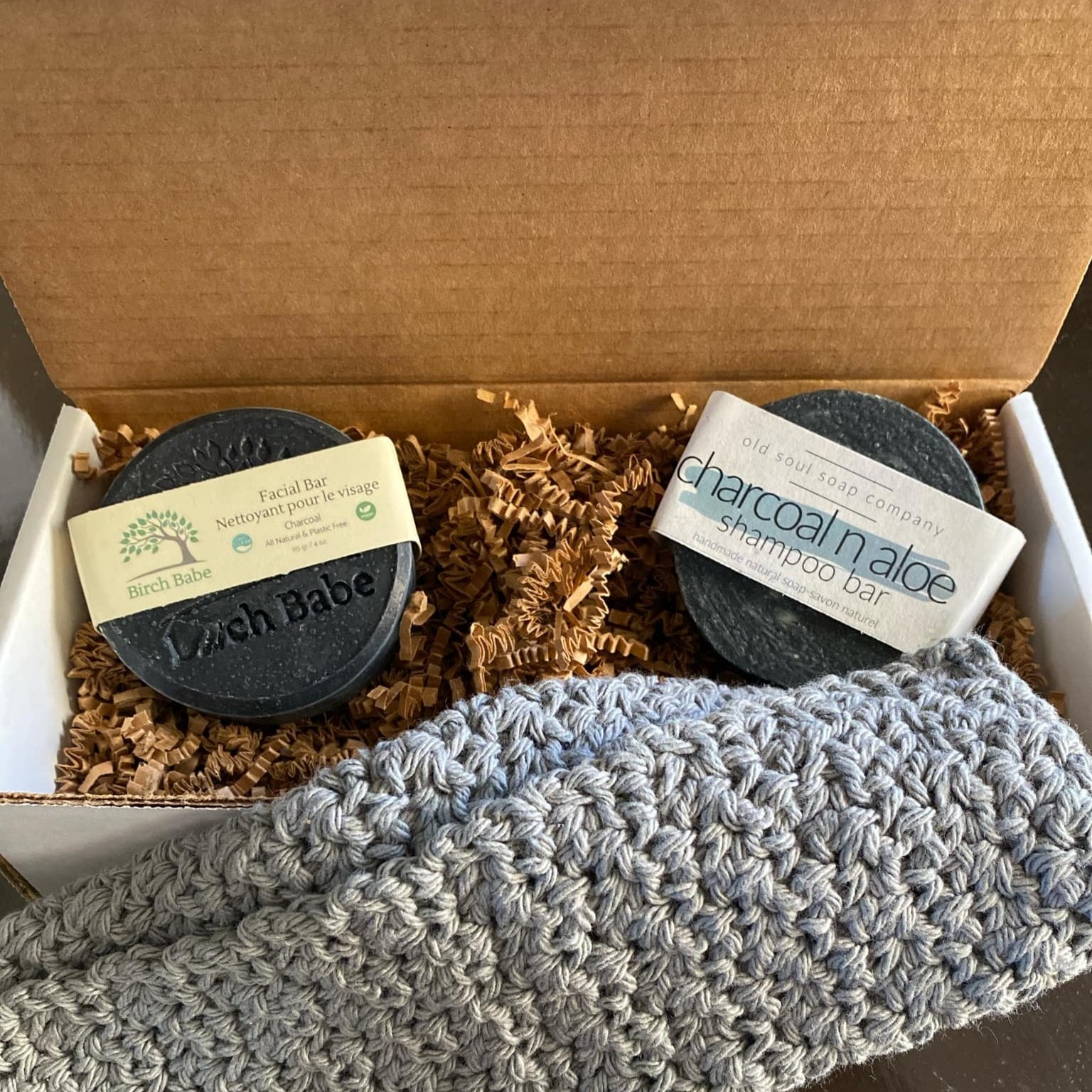 This activated charcoal gift set features three Canadian made products a round Birch Babe facial bar made with activated charcoal, a round charcoal 'n aloe shampoo bar from the Old Soul Soap Company and a handcrafted crocheted spa cloth in 100% cotton. The two labelled bars sit in a an open gift box on top of brown crinkle paper packaging with the spa cloth in the foreground. 