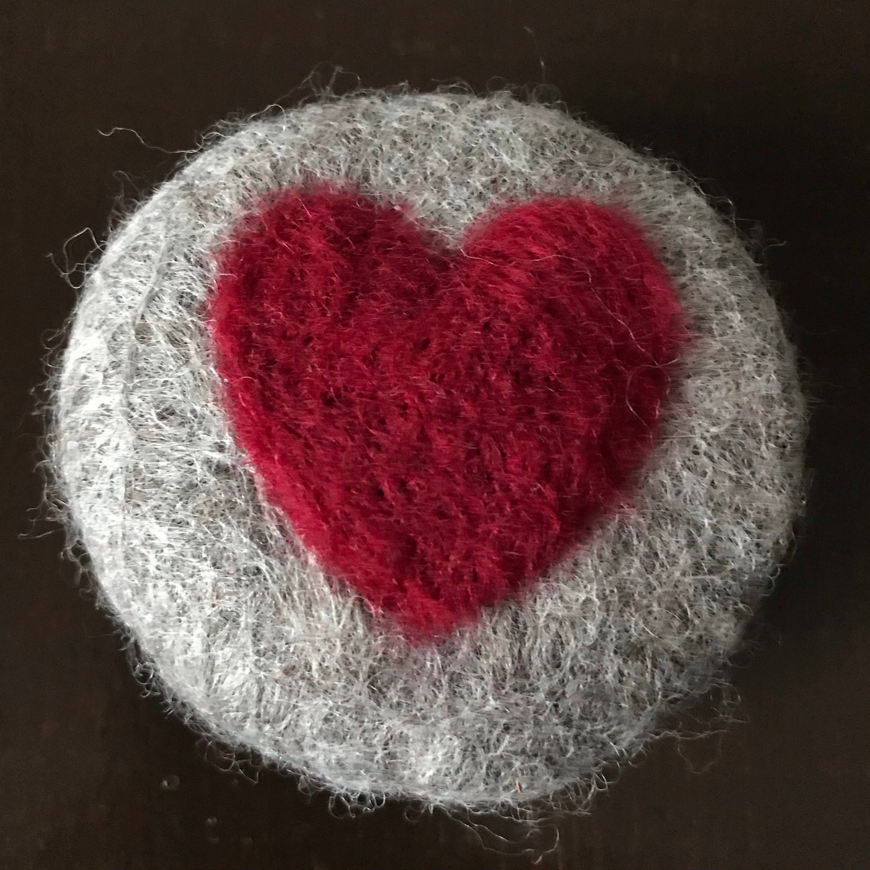 Round hand felted essential oil apple cinnamon scented soap made with Ontario cider and 100 per cent charcoal grey wool with a needle felted heart in the middle is pictured on a table. 