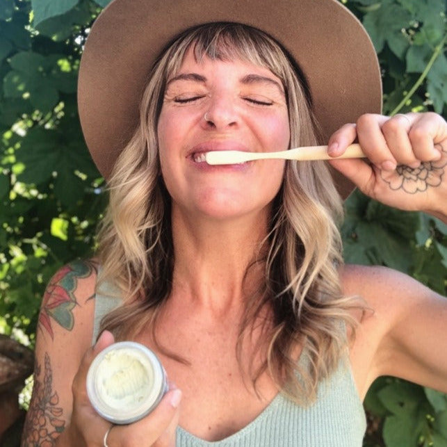 Smiling woman brushing teeth with Nelson Naturals Moringa Toothpaste in a jar using a bamboo toothbrush, promoting natural and sustainable oral care.