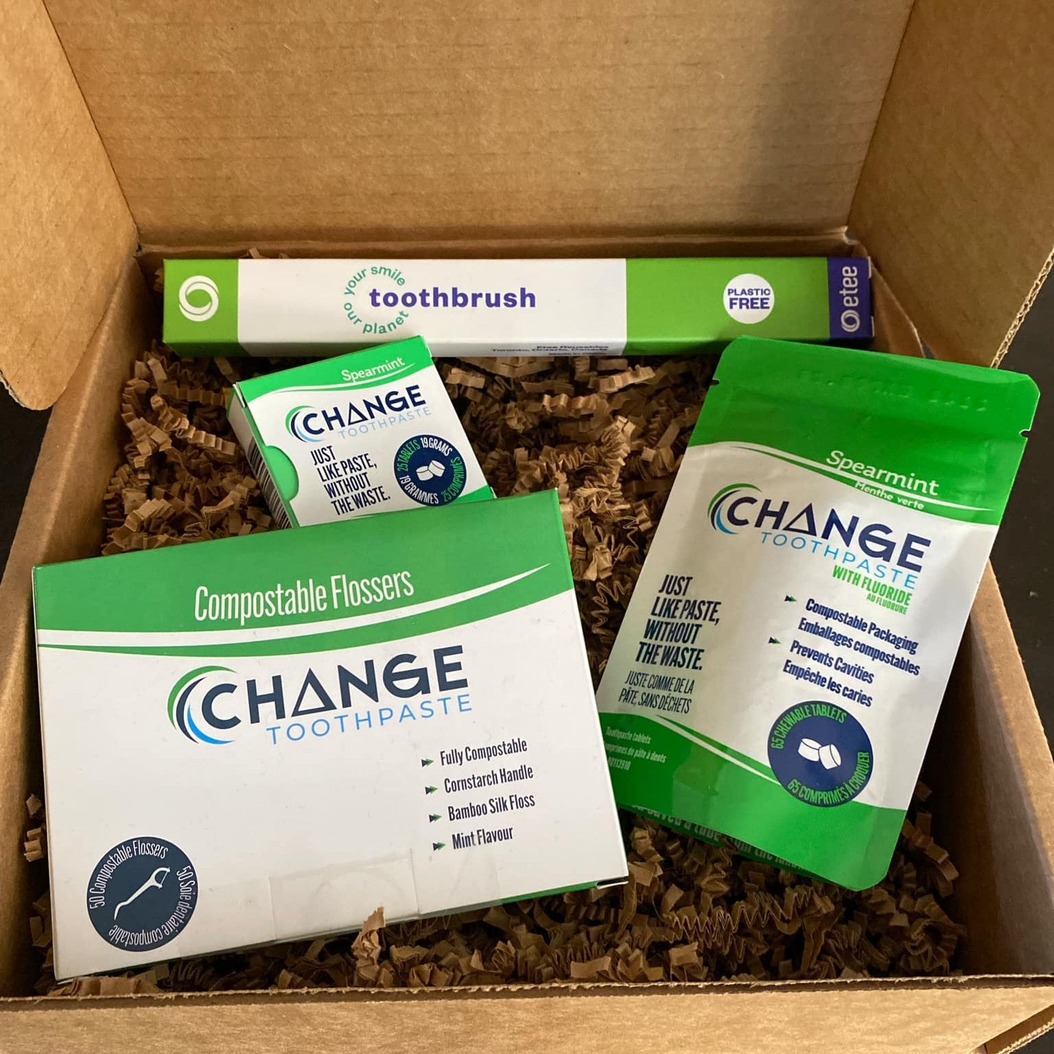 This dental care starter kit features three products from Change toothpaste. A package of 50 compostable flossers, a 65 tablet pouch of spearmint toothpaste tablets with fluoride, a tin of 25 regular spearmint toothpaste tablets and a bamboo toothbrush with a replaceable head free etee. These items are shown in an open gift box on top of brown crinkle paper packaging.