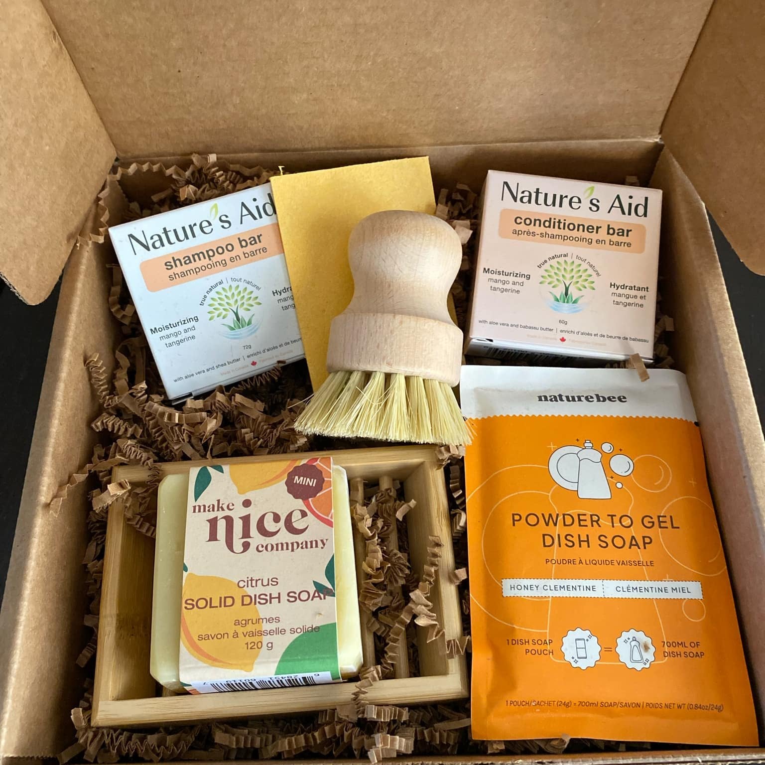 This zero-waste Citrus Gift Box of essentials includes Mango Tangerine shampoo and conditioner bars from Nature’s Aid, a Mini Citrus Solid Dish Soap Bar from the Make Nice Company, Honey Clementine Powder-to-Gel Dish Soap from Nature Bee, a biodegradable cellulose sponge, a bamboo soap tray, and a durable pot scrubber. These items are shown in an open gift box with the products sitting on brown crinkle paper packaging.