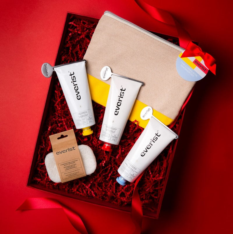 For holiday gift set display purposes, a full-size Shampoo Concentrate tube, full-size Deep Conditioning Concentrate tube, full-size Body Wash Concentrate tube, the Compostable Konjac Sponge and Everist Travel Bag  are displayed in a red gift box with red ribbon and red crinkle paper on a red background.