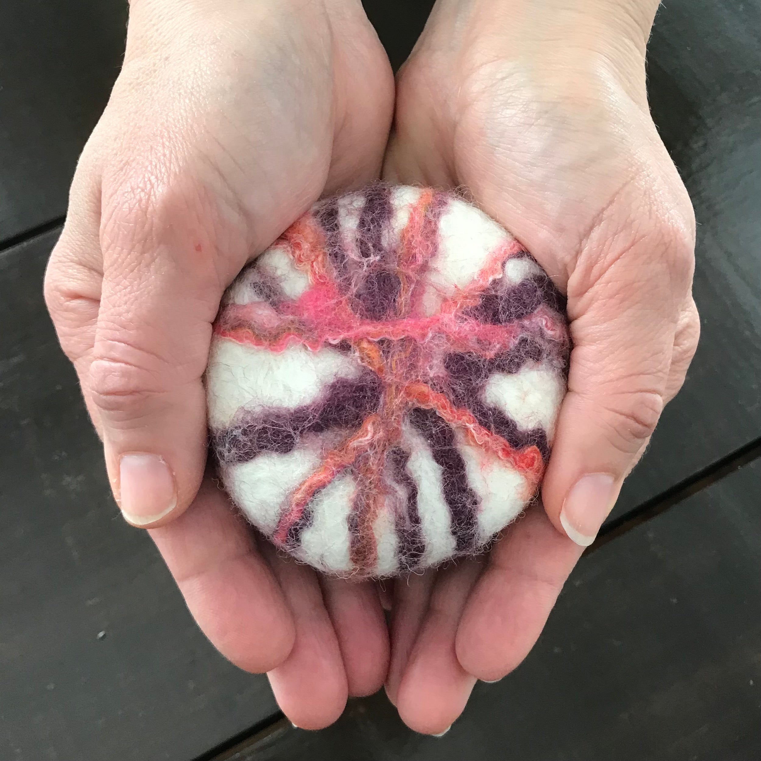 A natural essential oil floral sage scented felted soap with a purple, pink and orange felt pattern on a white background  made in Canada out of  100 per cent wool is held in a woman's palm