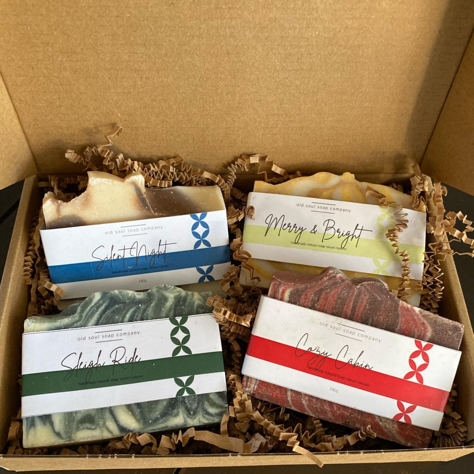 Four labelled handcrafted Old Soul Soap Company 'Silent Night', 'Merry & Bright', 'Sleigh Ride' and 'Cozy Cabin'  bar soaps in an open brown Kraft gift box sitting on brown crinkle paper packaging. 