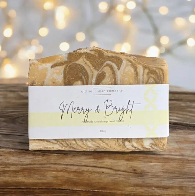 Light yellow and white labelled Merry and Bright Canadian made Old Soul Soap Company bar soap with yellow white and gold swirls with a Frankincense and peppermint scent sits on a wood table with white lights in the background.