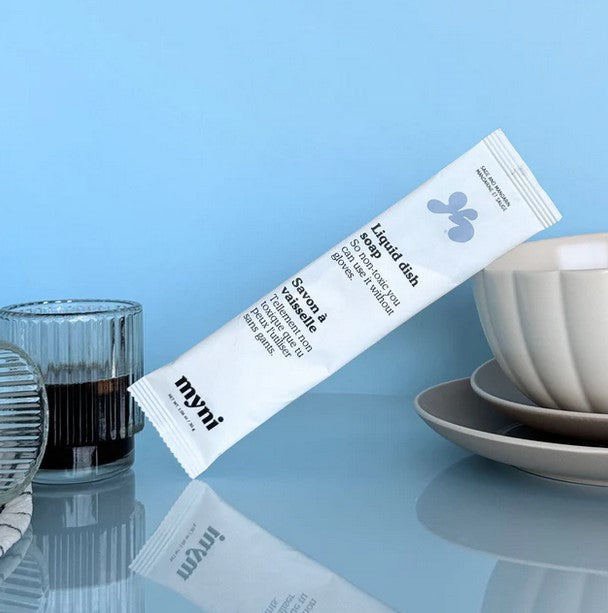 A compostable pouch of myni dish soap concentrate powder sits amid some clean dishes on a blue background.