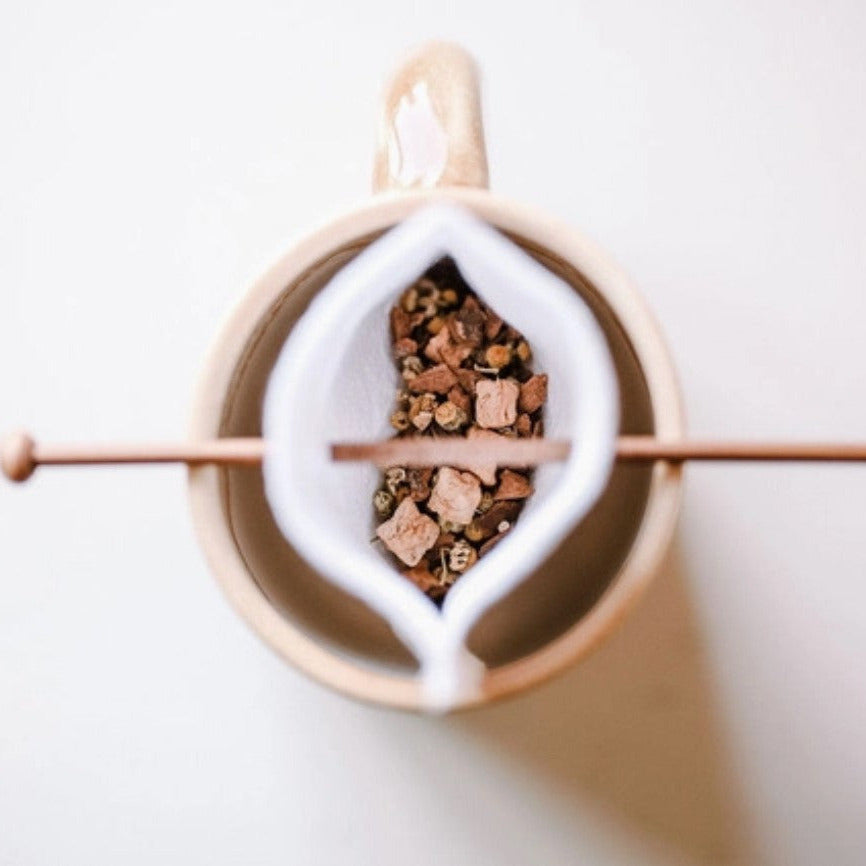 Top view of reusable organic cotton tea bag filled with loose leaf tea and wooden stick resting over mug – plastic-free tea brewing alternative.