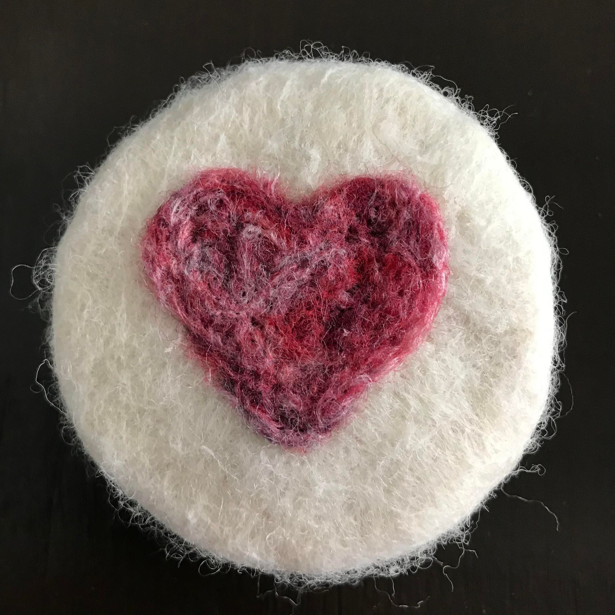 A round hand felted rose geranium essential oil  soap made in Canada in 100% white wool with a red textured heart on the top sits on a brown table