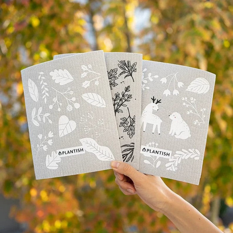 Hand holding Into the Woods Swedish Sponge Cloth Set of 3 by Plantish – grey Swedish dishcloths with First Frost, Herbal Remedy, and Deer Bear designs inspired by wildcrafting, with fall foliage in the background.