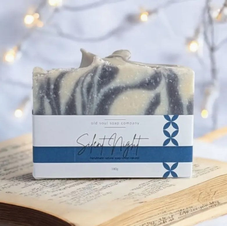 A blue and white labelled Silent Night Canadian made Old Soul Soap Company bar soap with swirls and a refreshing tangerine, bergamot lime, pine and wintergreen scent sits on a  stack of vintage paper with white lights in the background.