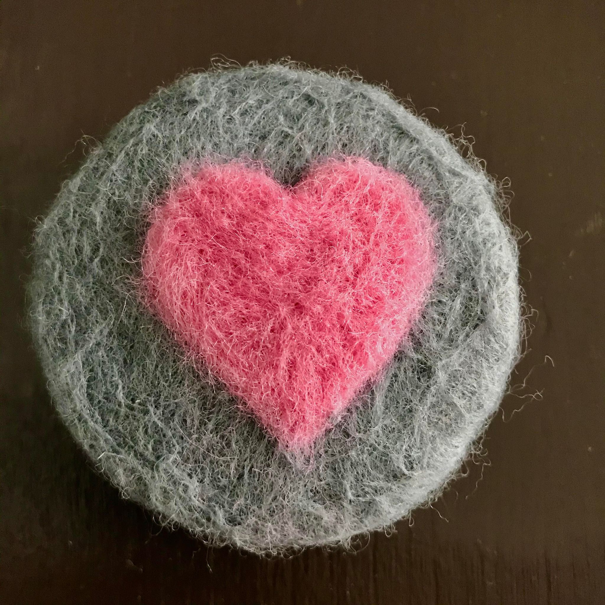 A round hand felted rosemary mint essential oil  soap made in Canada in 100% grey with a pink heart on the top sits on a brown table