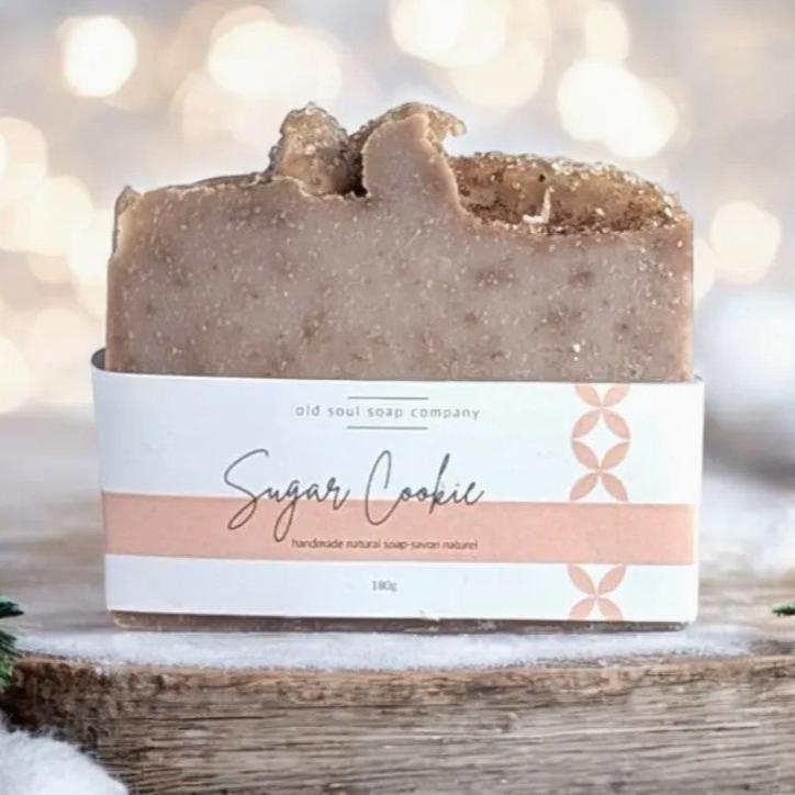 A labelled Sugar Cookie Canadian made Old Soul Soap Company bar soap with a sweet fragrant scents sits on a wood table with white lights in the background.