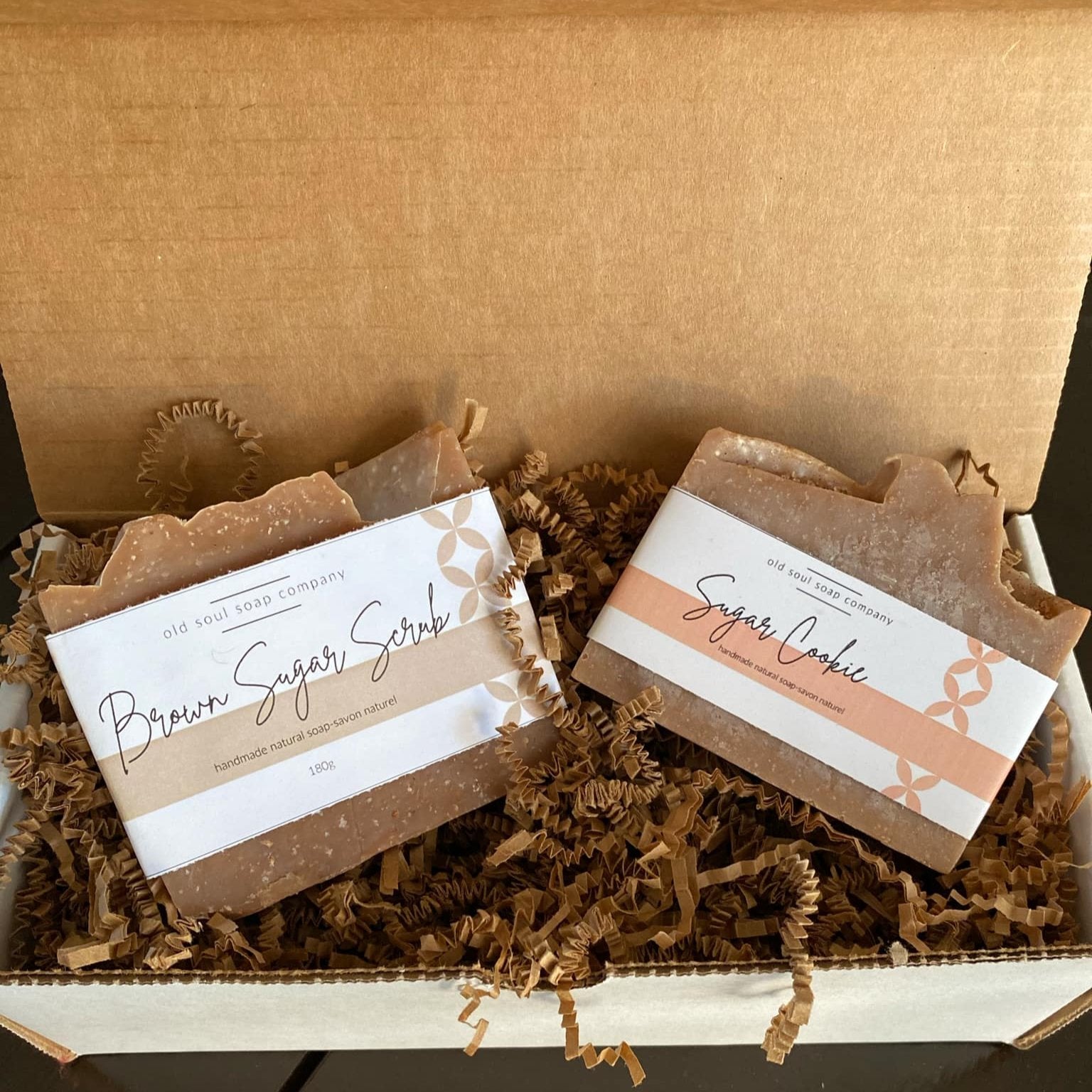 Two labelled handcrafted Old Soul Soap Company 'Brown Sugar Scrub' and 'Sugar Cookie' bar soaps in an open white gift box sitting on brown  crinkle paper packing. 