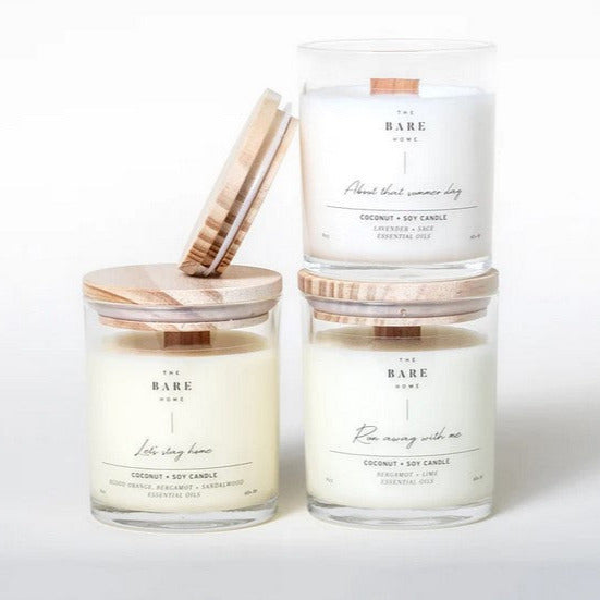 Three candles in clear glass jars with wood tops on a white background. About That Summer Day (Lavender + Sage) has the lid off and sits on top of the other two candles -  Let’s Stay Home (Blood Orange, Bergamot + Sandalwood) and Run Away With Me (Bergamot + Lime). Candles are all scented with essential oils.