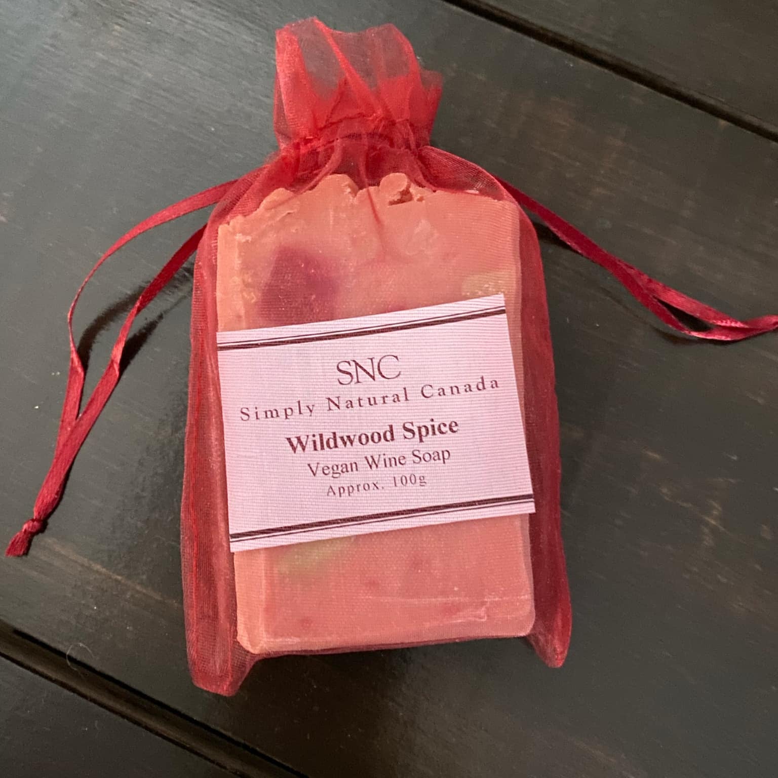 A labelled Wildwood Spice vegan wine soap handcrafted in Canada by SNC in a red organza bag sitting on a table.