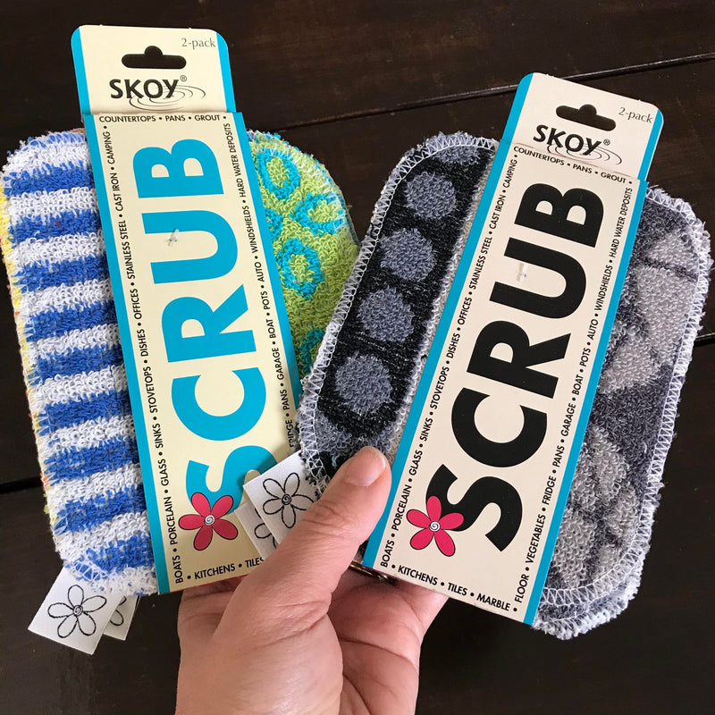A hand is holding two Skoy Scrub Cloth sets of two, the one on the left features assorted colors and the one on the right is monochromatic in black and grey