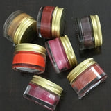 seven shades of  lip and cheek tint made in canada by birch babe in absolutely fabulous (pink), backcountry diva (medium red), fireside temptation (dark purpleish red), ruby rebel (ruby red), sunset seeker (orange red), vacation mode with sun protection (copper gold), and vintage rose (dusty pink) made by birch babe and sold in 20 ml glass jars