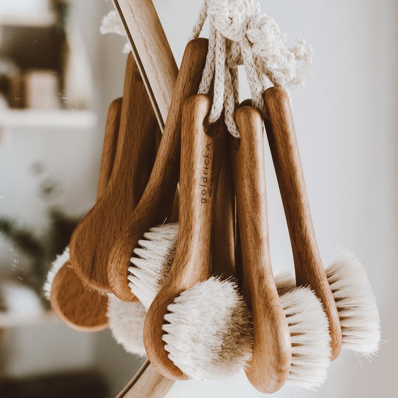 A bunch of Goldrick Natural Living wooden face brushes hanging beside a mirror
