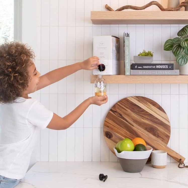 Child refilling a blood orange, bergamot and sandalwood glass refill bottle with a 3 L refill box sitting on a kitchen shelf