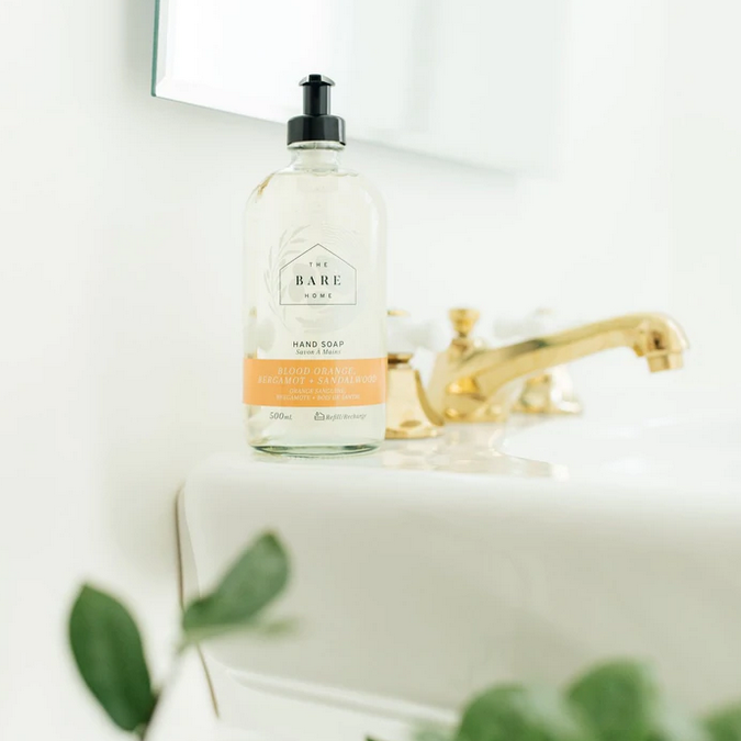 Canadian made The Bare Home Blood Orange, Bergamot Lime and Sandalwood essential oil scented natural hand soap in a 476 ml refillable glass pump bottle sits beside a polished bathroom faucet with plants in the foreground 